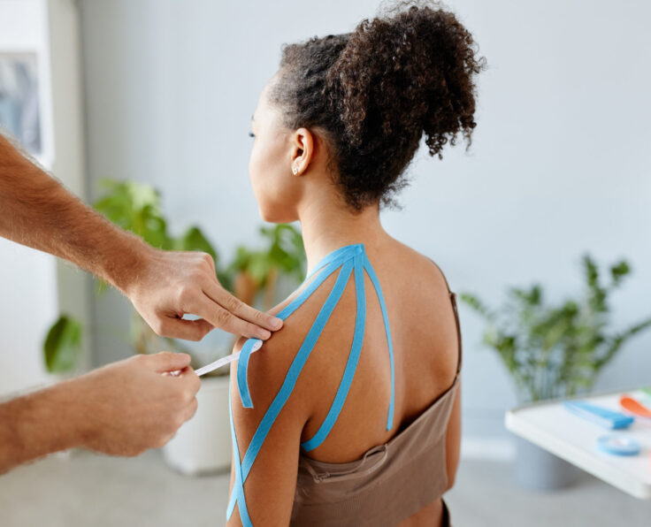Close up rehabilitation therapist putting k tape on shoulder of young African American woman in physiotherapy clinic